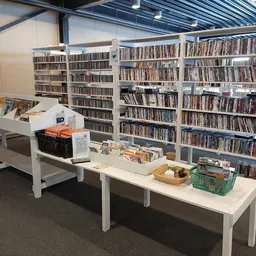 Interieur van kringloopwinkel met witte tafels en volle cd/dvd-rekken op de achtergrond