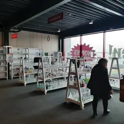 Winkelvloer met witte stellingen vol spullen, boekenrek links, grote ramen met uitzicht, bezoeker loopt door de winkel