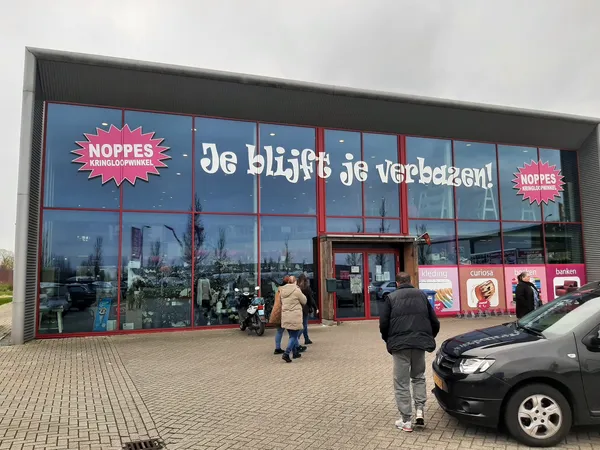 Gevel van kringloopwinkel met grote ramen, rode deur en rode NOPPES-logo's op blauwe achtergrond