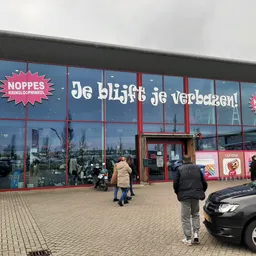 Gevel van kringloopwinkel met grote ramen, rode deur en rode NOPPES-logo's op blauwe achtergrond