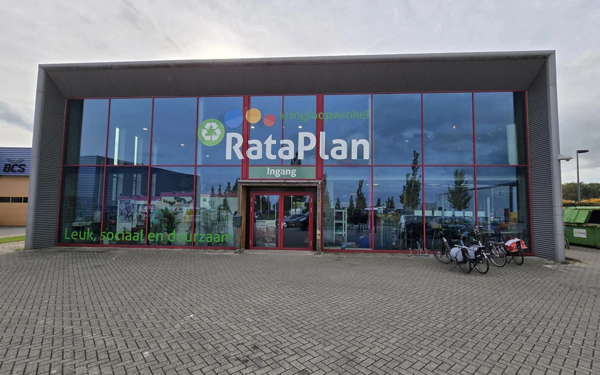 Moderne gevel van kringloopwinkel Rataplan met grote glazen pui en rode deur, gelegen aan straatzijde met parkeerplaats.