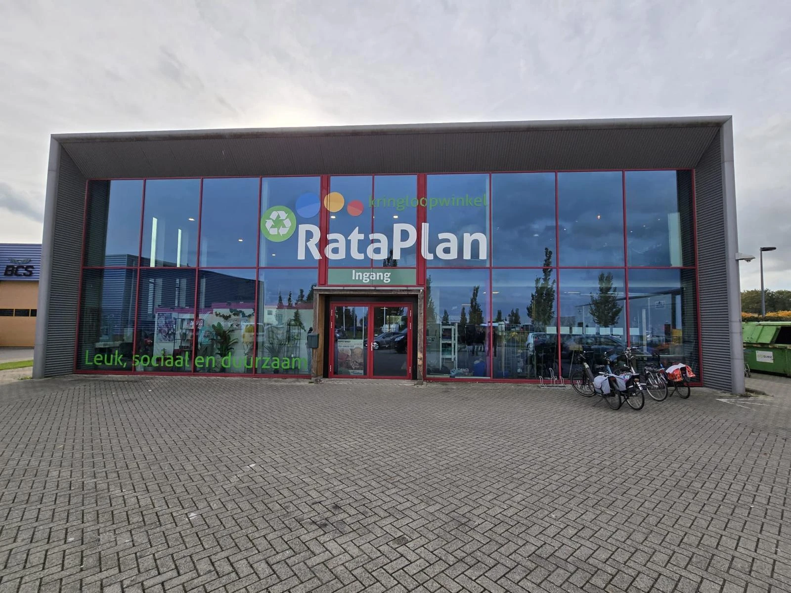 Moderne gevel van kringloopwinkel Rataplan met grote glazen pui en rode deur, gelegen aan straatzijde met parkeerplaats.