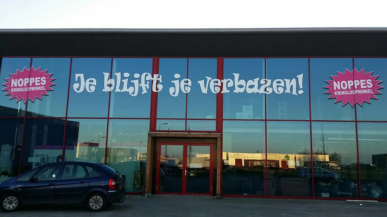 Glazen gevel van kringloopwinkel met rode stalen raamkozijnen en witte tekst 'Je blijft je verbazen!' op blauw glas