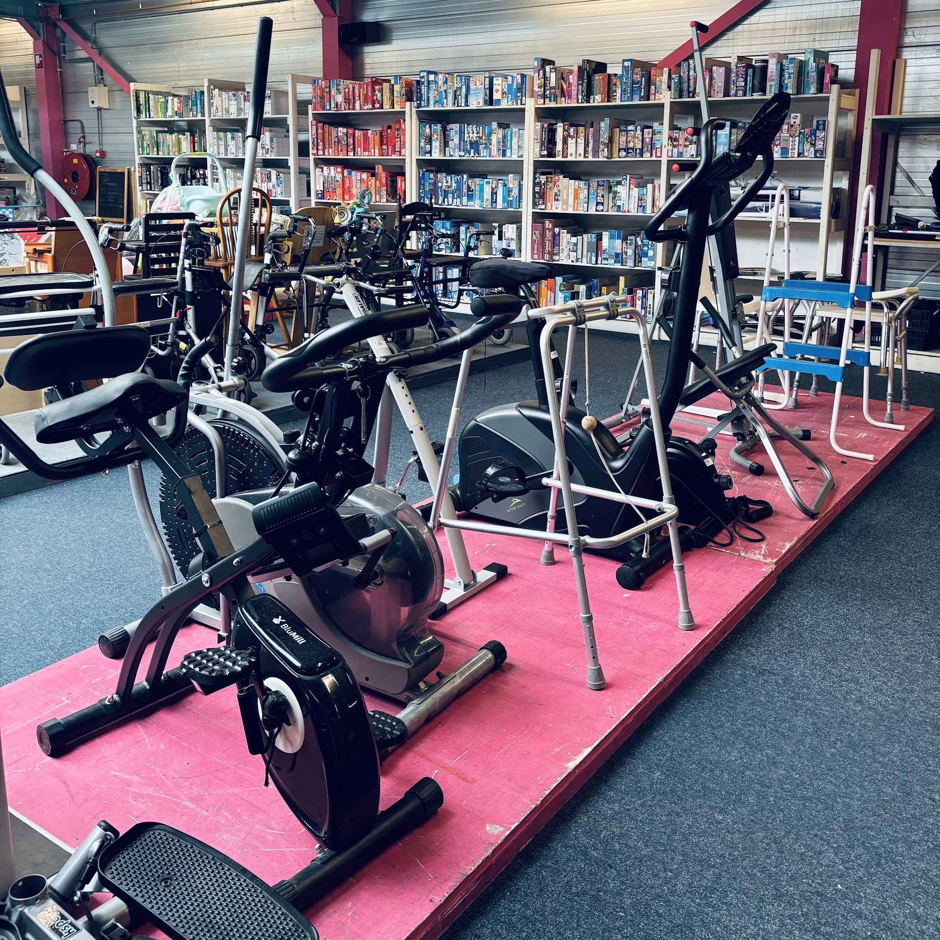 Fitnessapparaten en hometrainers op roze mat in kringloopwinkel, boekenkasten op achtergrond