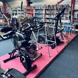 Fitnessapparaten en hometrainers op roze mat in kringloopwinkel, boekenkasten op achtergrond