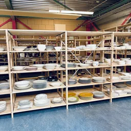 Houten stellingen met uitgestalde serviesgoed: stapels borden, schalen en ander tafelservies in wit, blauw en geel