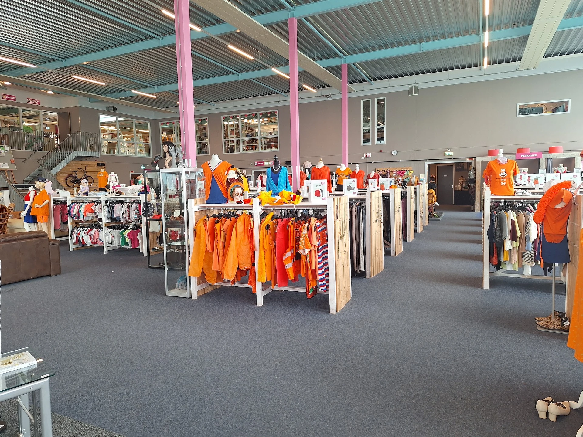 Ruim winkelbinnenruimte met kledingrekken vol oranje kleding, roze zuilen en grijs plafond