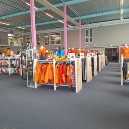 Ruim winkelbinnenruimte met kledingrekken vol oranje kleding, roze zuilen en grijs plafond