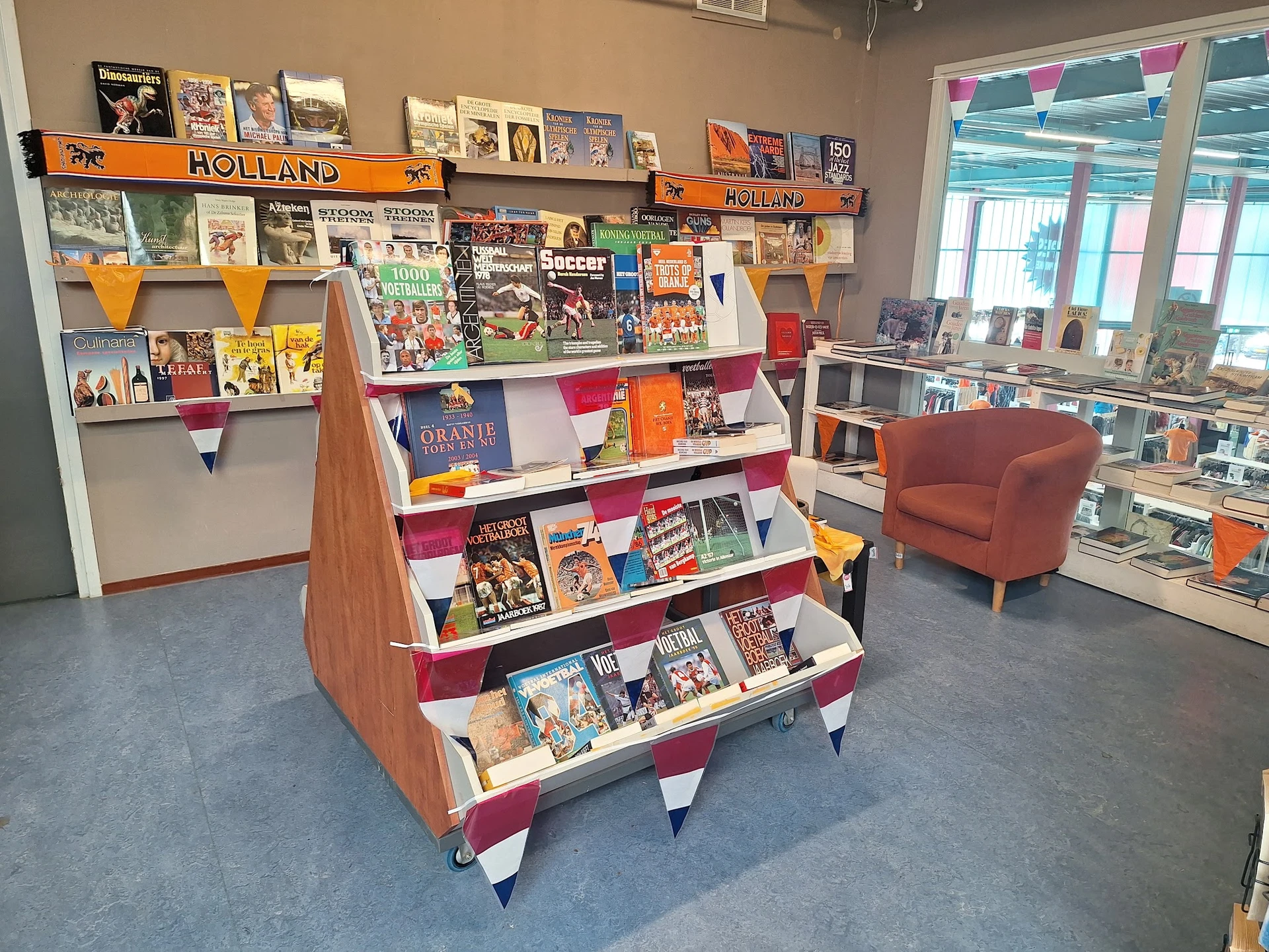 Winkelinterieur met boekenrek op wielen en wandkasten vol boeken en posters, oranje stoeltje op de achtergrond