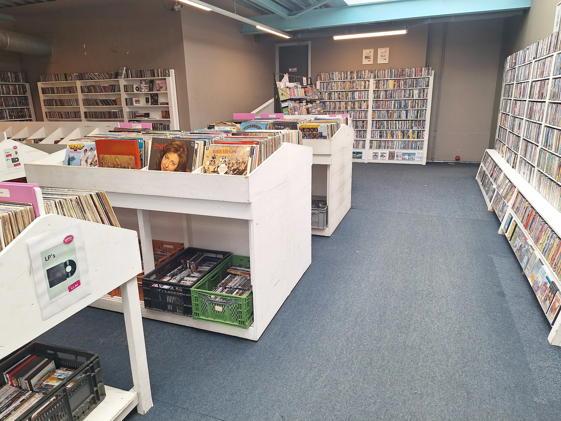 Binnenaanzicht van kringloopwinkel met stellingen vol cd's, dvd's en boeken, witte kastenen display in het midden