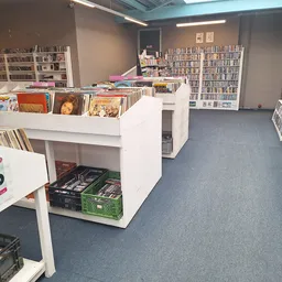 Binnenaanzicht van kringloopwinkel met stellingen vol cd's, dvd's en boeken, witte kastenen display in het midden