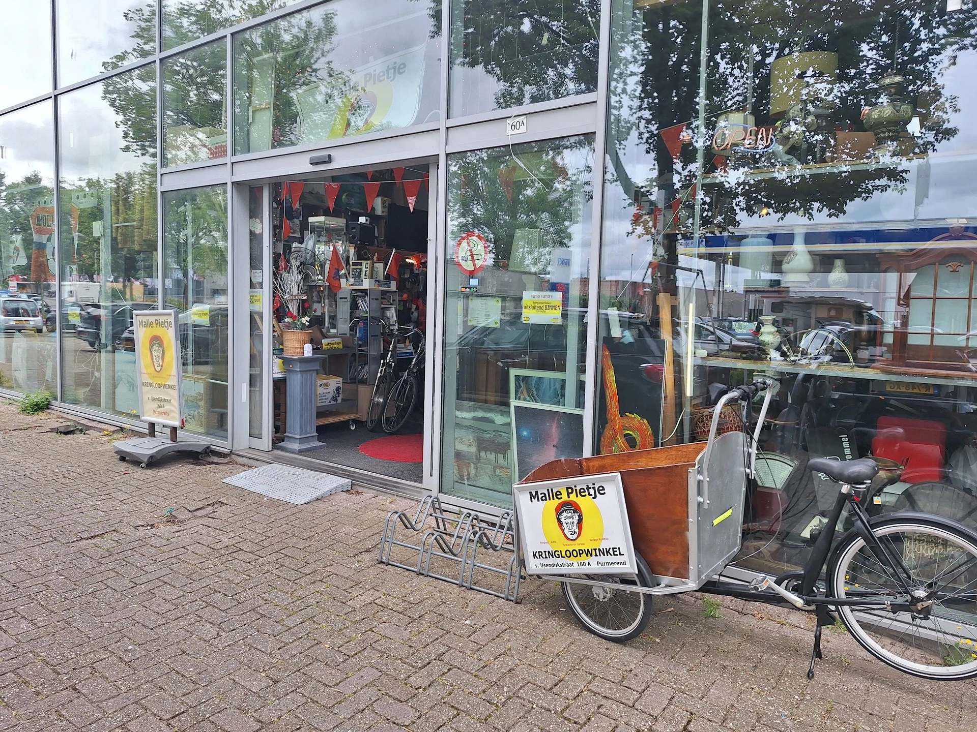 Voorkant van kringloopwinkel met grote glazen pui, fietsen ernaast geparkeerd en een geel bord met Malle Pietje logo
