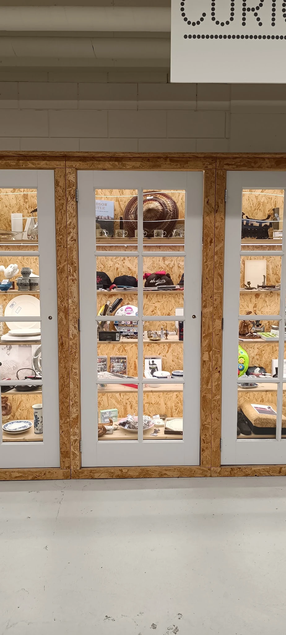 Glazen vitrinekasten met kurken wanden en witte raamprofielen, gevuld met diverse spullen zoals servies, hoeden en decoratiev