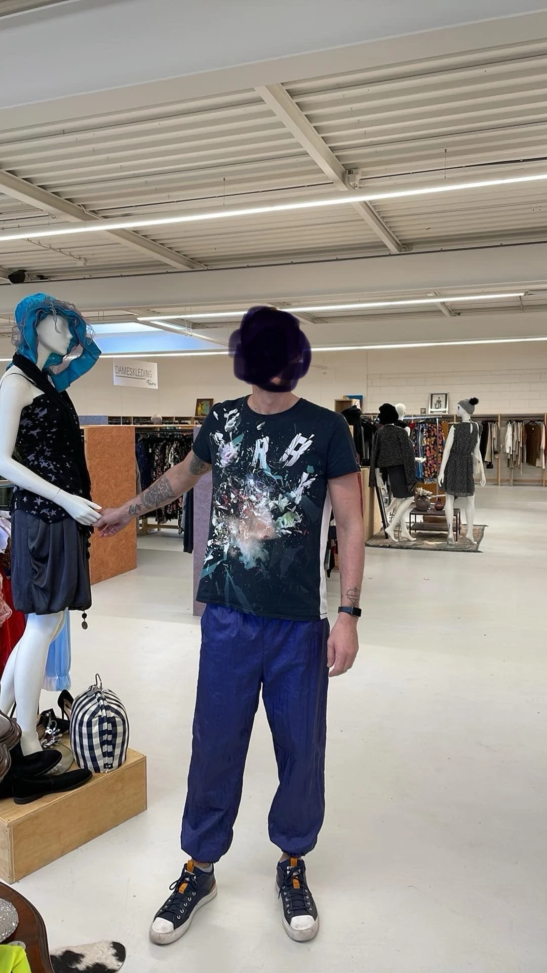 Mannequin met blauwe pruik en pop-up klant in grijs t-shirt in kringloopwinkel met kledingrekken op achtergrond