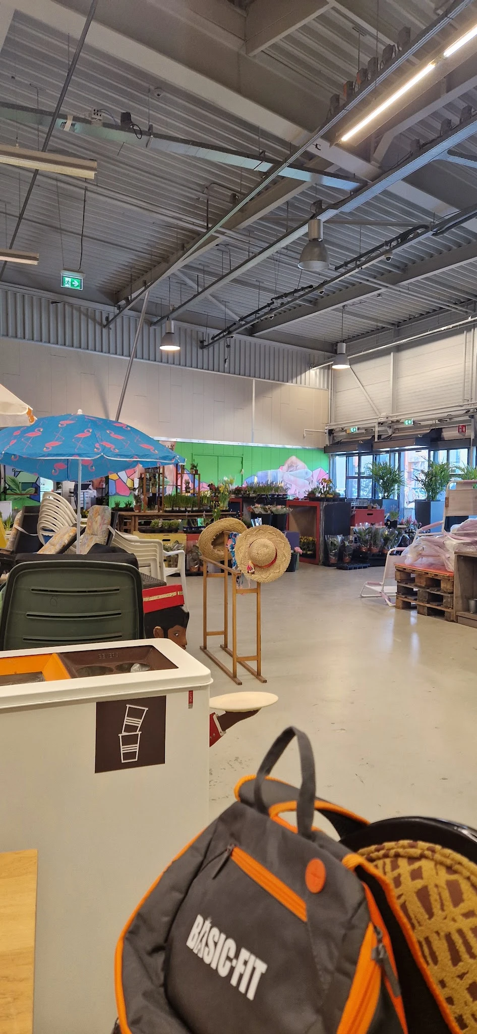 Winkelvloer van kringloopwinkel met diverse spullen: parasol, strohoedenrek, groene lade, stellingen met plantenpotten en hui