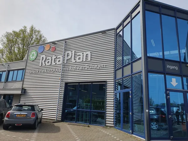 Gevel van kringloopwinkel RataPlan Schagen met naam leesbaar op de voorgevel, glazen deuren, parkeerplaats met auto's