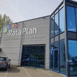 Gevel van kringloopwinkel RataPlan Schagen met naam leesbaar op de voorgevel, glazen deuren, parkeerplaats met auto's