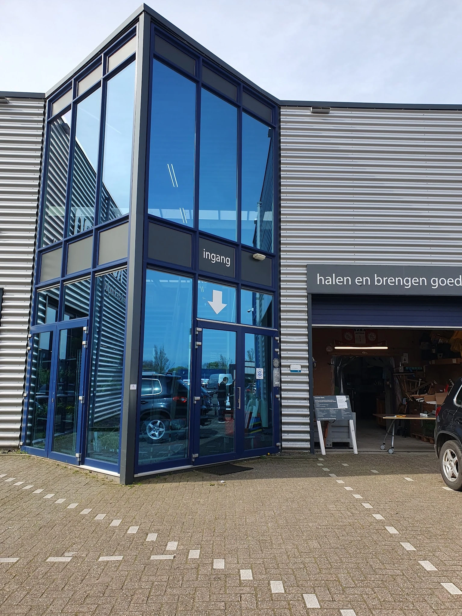 Glazen ingangsdeuren van kringloopwinkel met 'Ingang' en pijl aangegeven, naast winkelpui met tekst 'halen en brengen goed'