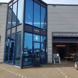 Glazen ingangsdeuren van kringloopwinkel met 'Ingang' en pijl aangegeven, naast winkelpui met tekst 'halen en brengen goed'