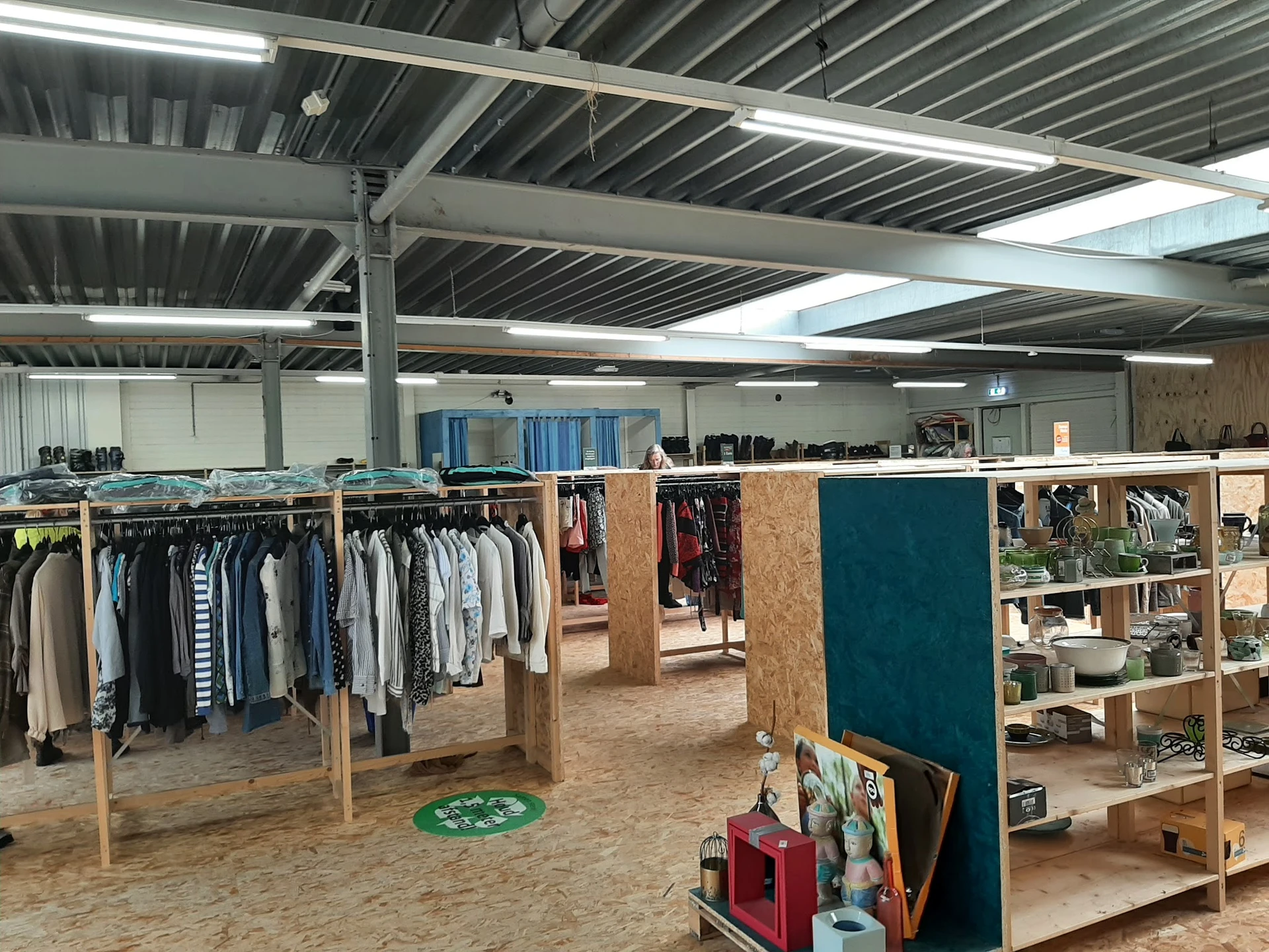 Ruime kringloopwinkel met kledingrekken, houten stellingen en dividers, industrieel plafond met TL-verlichting