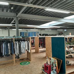 Ruime kringloopwinkel met kledingrekken, houten stellingen en dividers, industrieel plafond met TL-verlichting