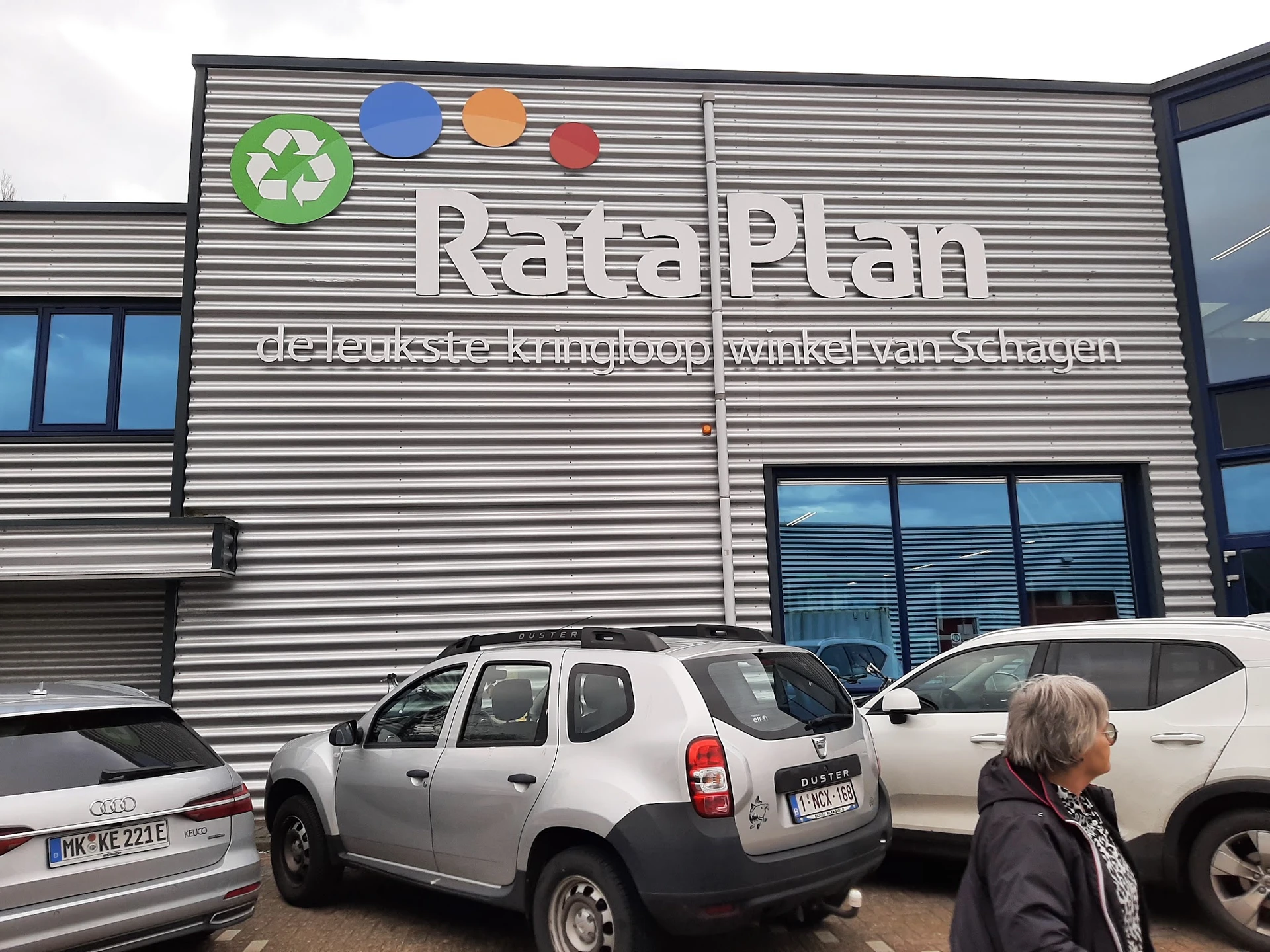 Gevel van kringloopwinkel RataPlan met recycling-logo en geparkeerde auto's ervoor