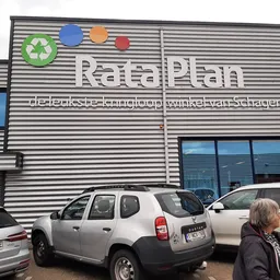 Gevel van kringloopwinkel RataPlan met recycling-logo en geparkeerde auto's ervoor