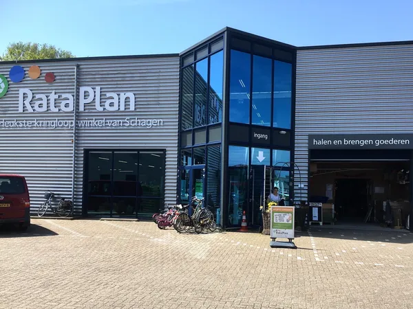 Gevel van kringloopwinkel RataPlan met grijze golfplaten, blauwe entree en fietsen geparkeerd ervoor