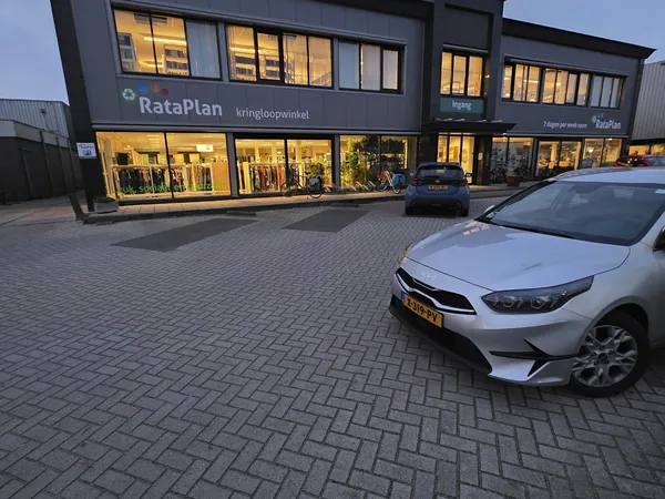 Gevel van kringloopwinkel RataPlan met verlichte etalage en parkeerplaats met twee auto's