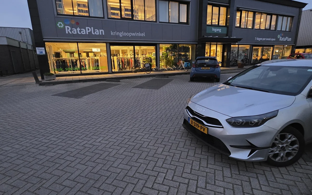 Gevel van kringloopwinkel RataPlan met verlichte etalages en geparkeerde auto's op het parkeerterrein