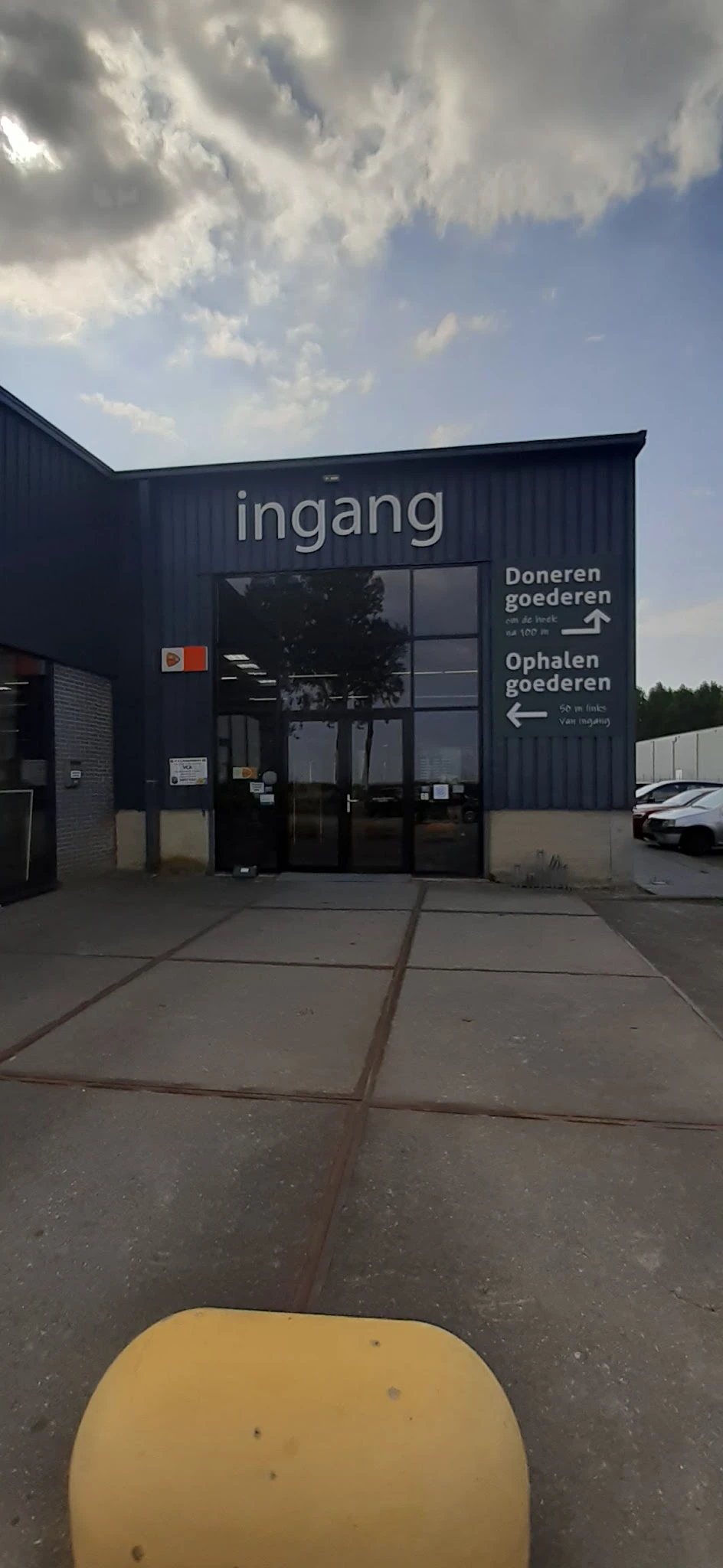 Ingang van kringloopwinkel met donkerblauw gevel, glazen deuren en wegwijzers voor doneren en ophalen