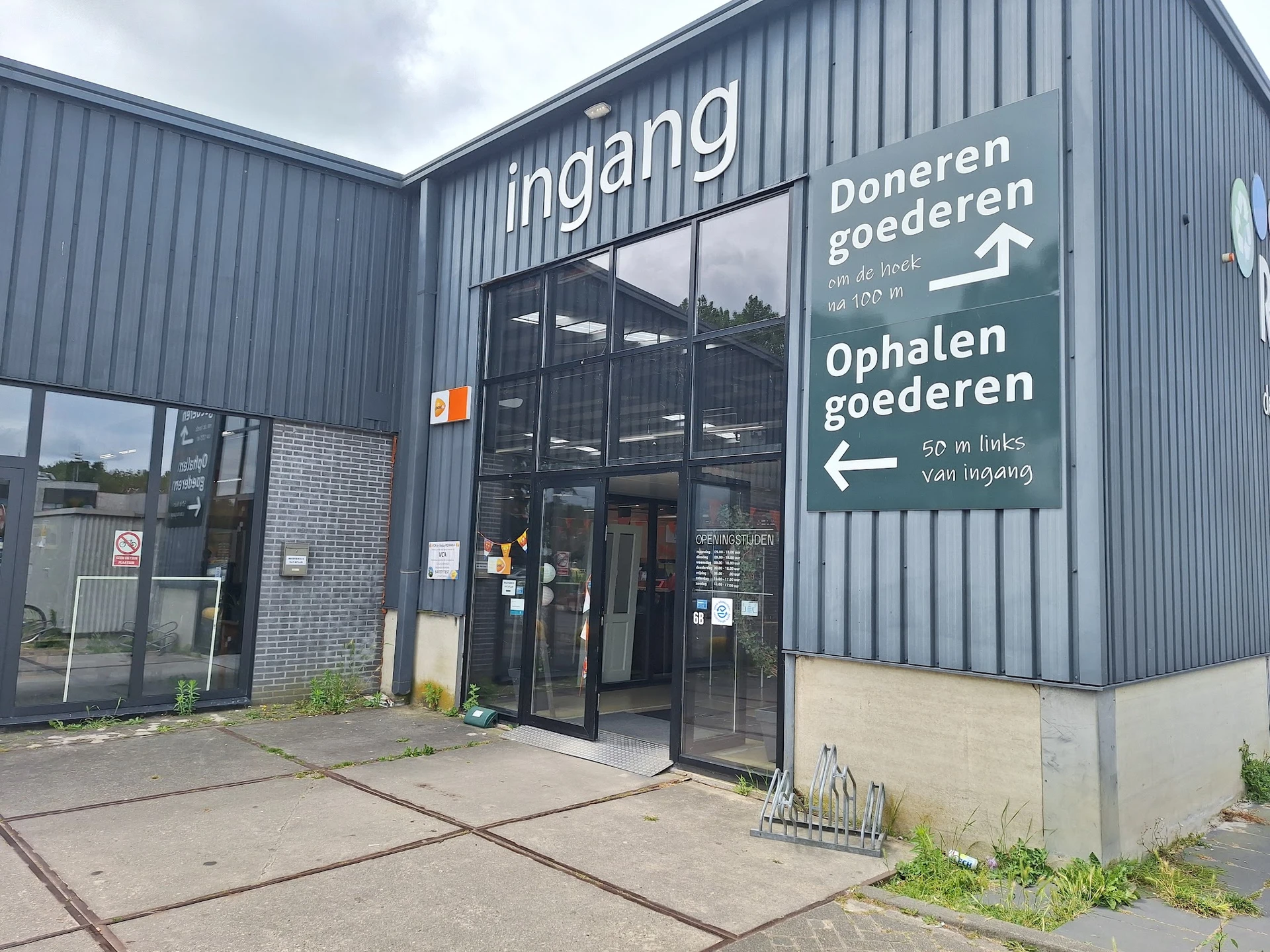 Grijze golfplaten gevel van kringloopwinkel met ingang, donkere ramen en witte richtingsborden voor doneren en ophalen