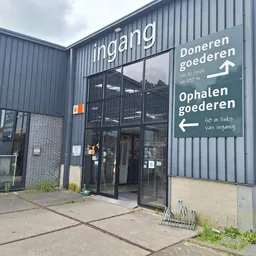 Grijze golfplaten gevel van kringloopwinkel met ingang, donkere ramen en witte richtingsborden voor doneren en ophalen