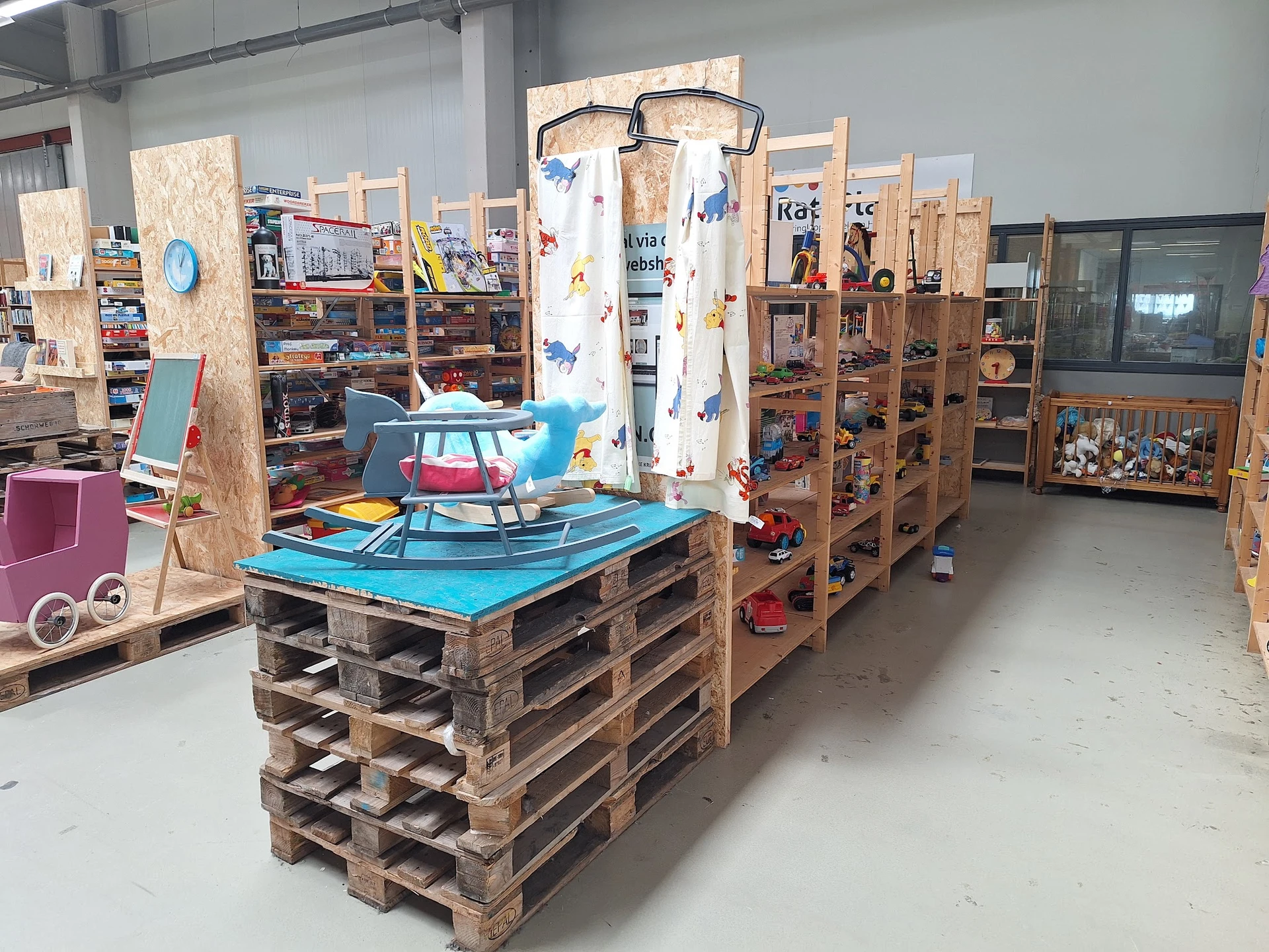 Winkelvloer met houten palletkasten, houten stellingen en speelgoed, waaronder een blauwe wipwagen onder een wit gordijn