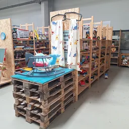 Winkelvloer met houten palletkasten, houten stellingen en speelgoed, waaronder een blauwe wipwagen onder een wit gordijn