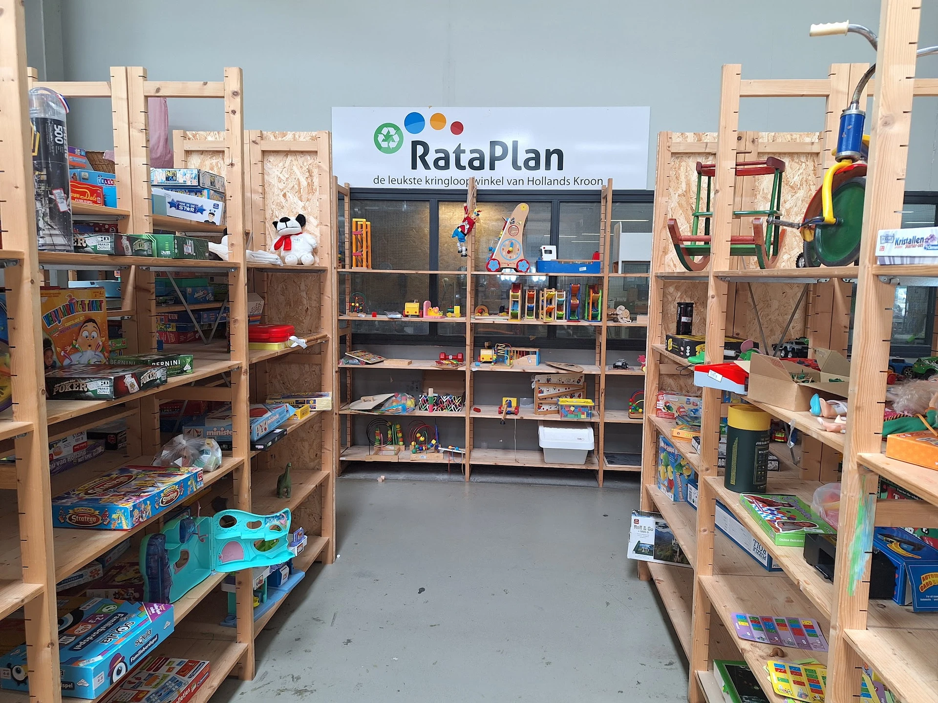 Winkelvloer met houten stellingen vol speelgoed, boeken en huishoudartikelen, RataPlan-bord zichtbaar