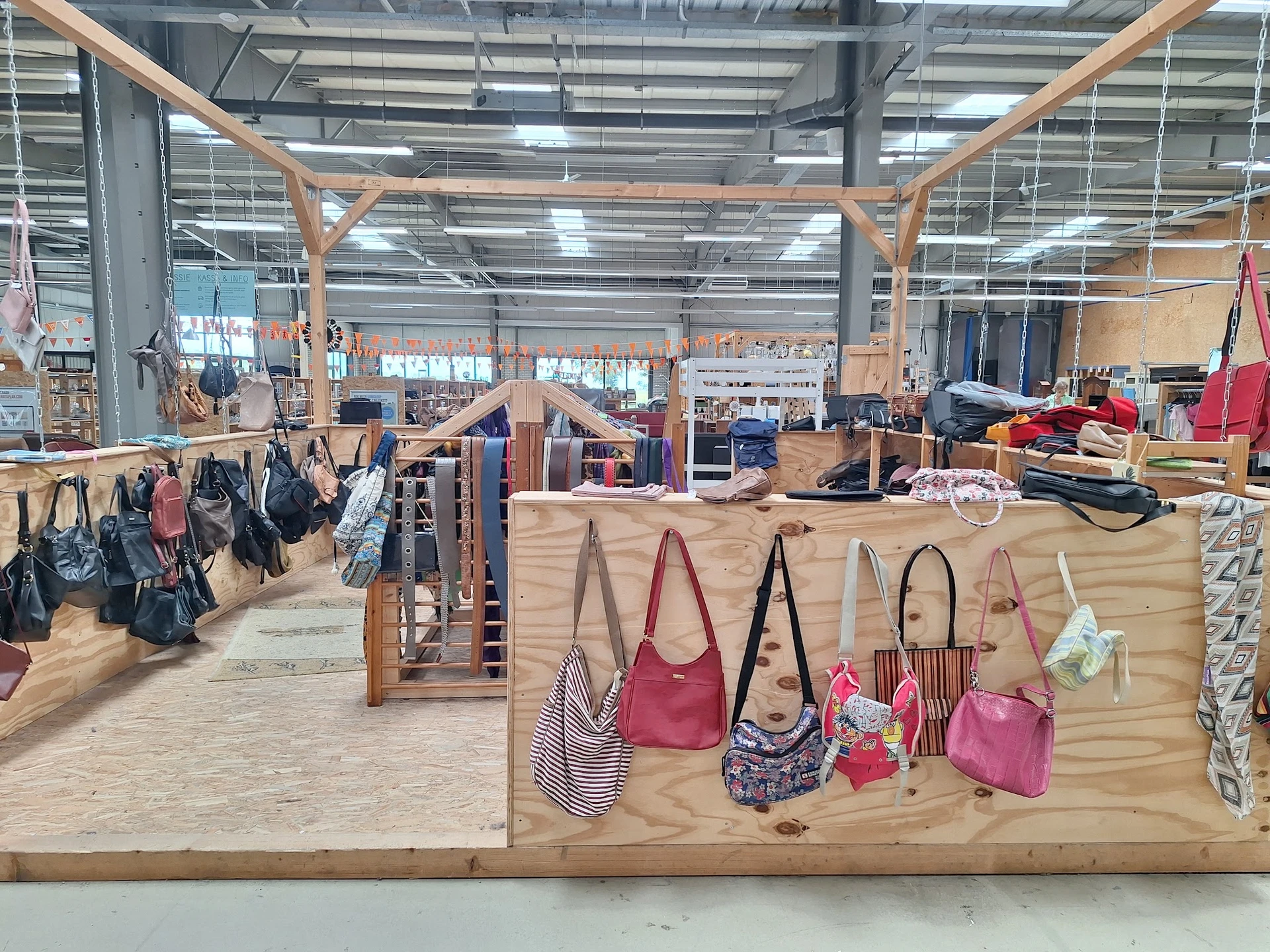 Uitgestalde tassen en handtassen aan houten display, met op de achtergrond meer tweedehands spullen in de kringloopwinkel