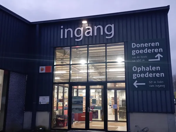 Avondfoto van de ingang van kringloopwinkel RataPlan met verlichte gevelreclame en bewegwijzering