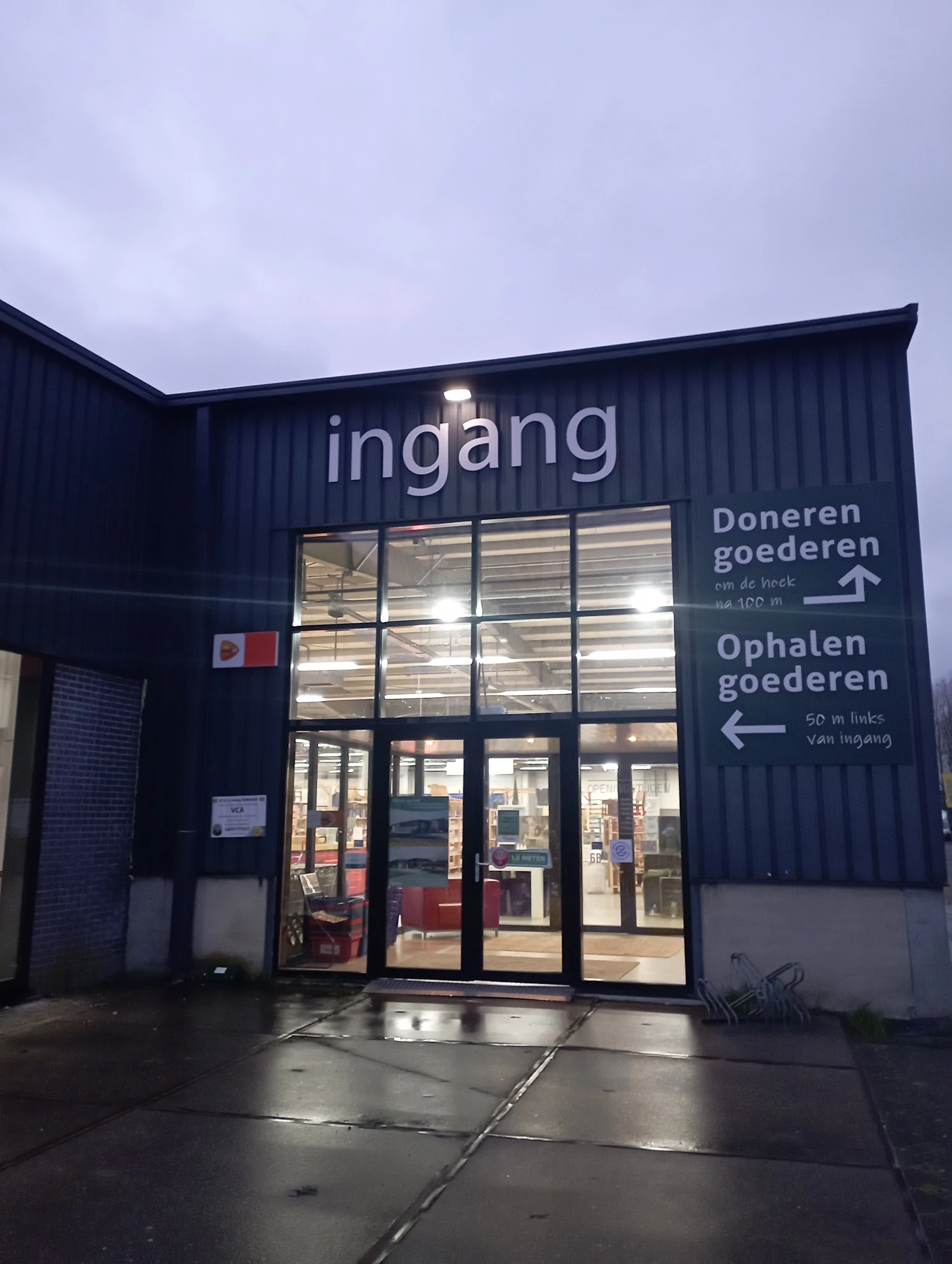 Avondfoto van de ingang van kringloopwinkel RataPlan met verlichte gevelreclame en bewegwijzering