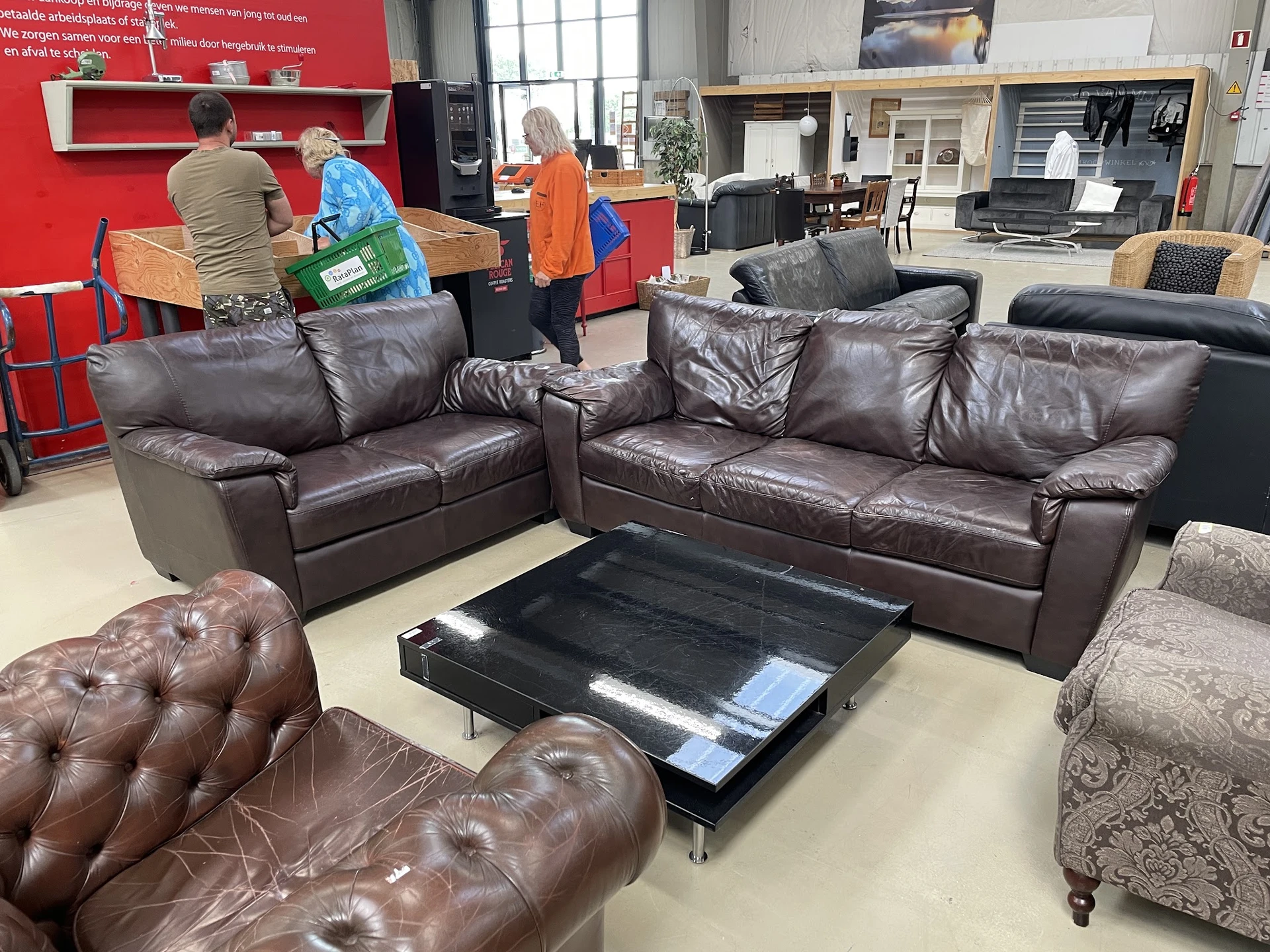 Bruine leren zitgroepen en fauteuils in een kringloopwinkel met rood accent op de achterwand