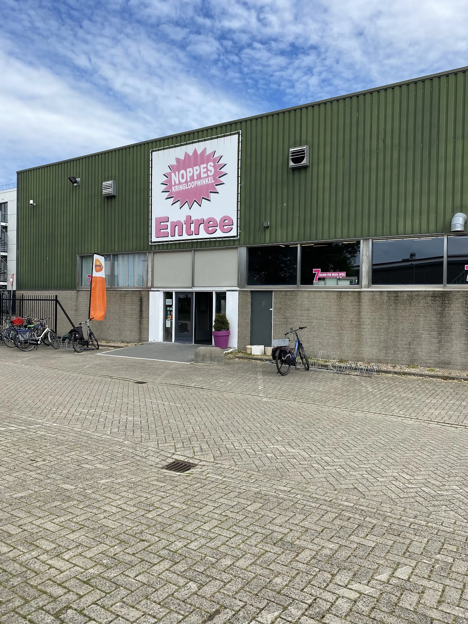 Gevel van kringloopwinkel met groen golfplaten dak, witte entree-bord met roze logo, en ingang op begane grond met fietsen er