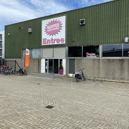Gevel van kringloopwinkel met groen golfplaten dak, witte entree-bord met roze logo, en ingang op begane grond met fietsen er