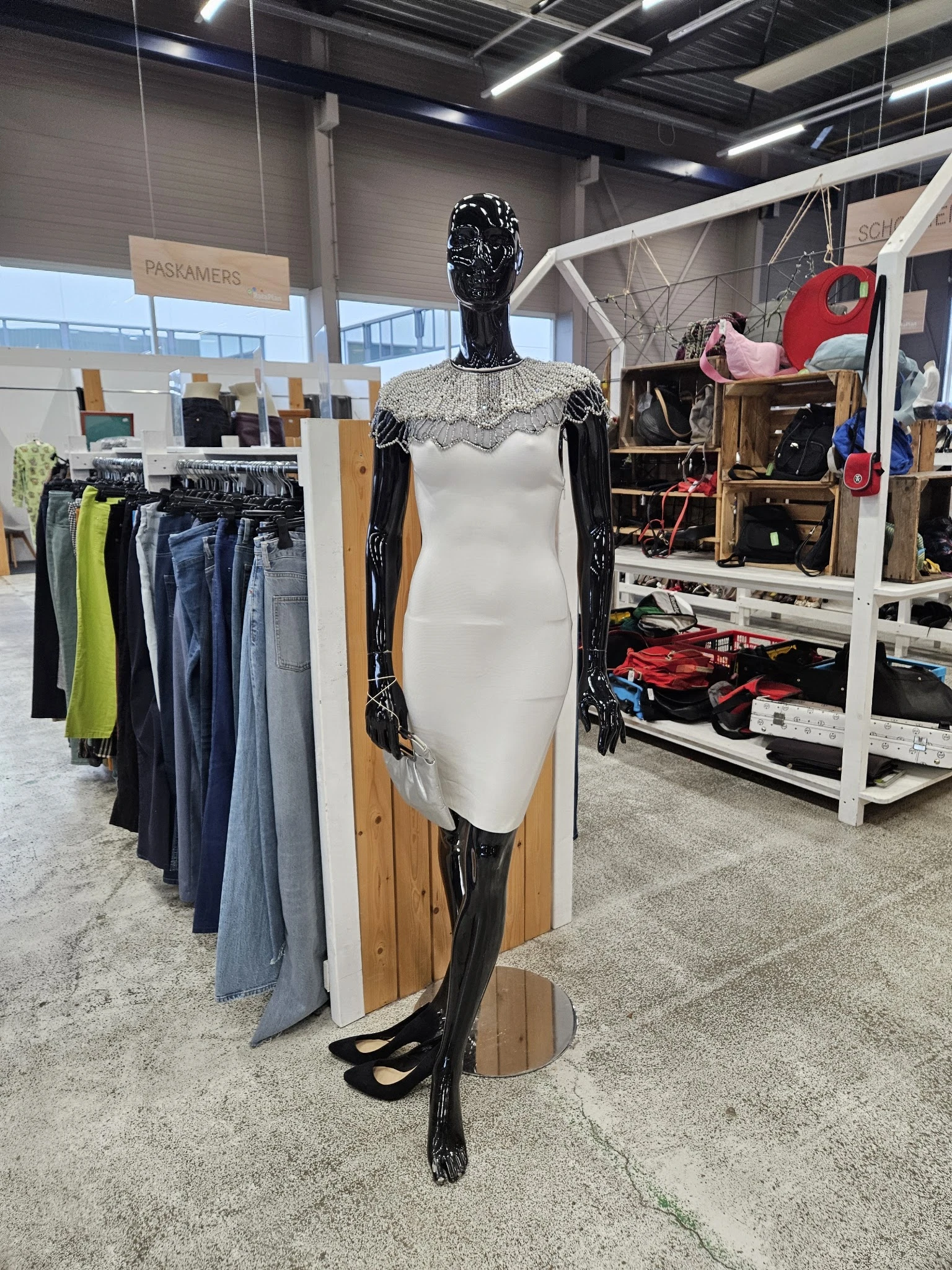 Etalagepop in witte jurk met zwarte mouwen en kantkraag, omringd door rekken met tweedehands kleding en andere winkelinventar