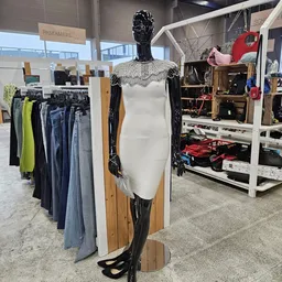 Etalagepop in witte jurk met zwarte mouwen en kantkraag, omringd door rekken met tweedehands kleding en andere winkelinventar