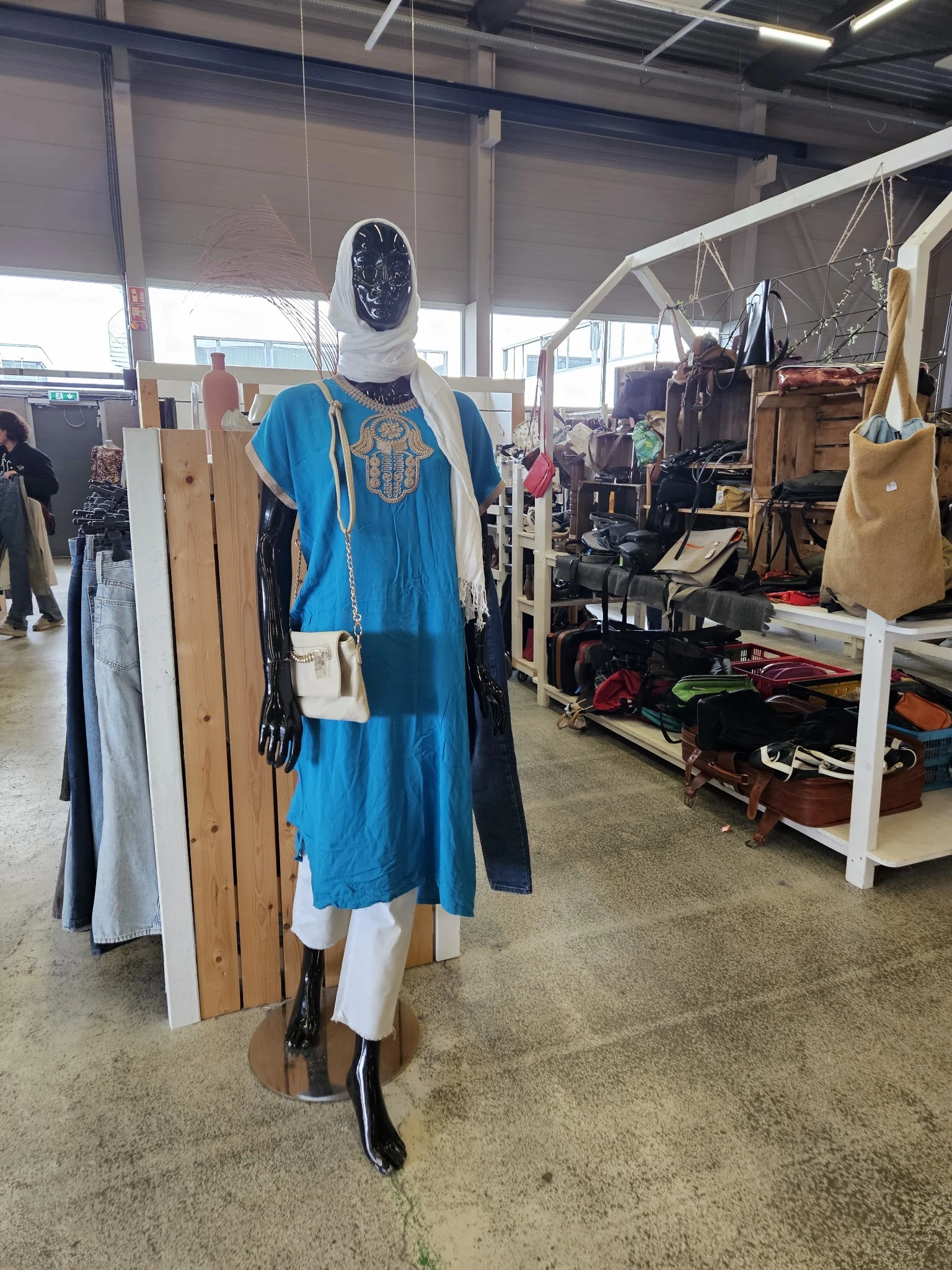 Etalage met tweedehands kleding op mannequin, waaronder een blauw kleed, en diverse kledingstukken op rekken en in bakken