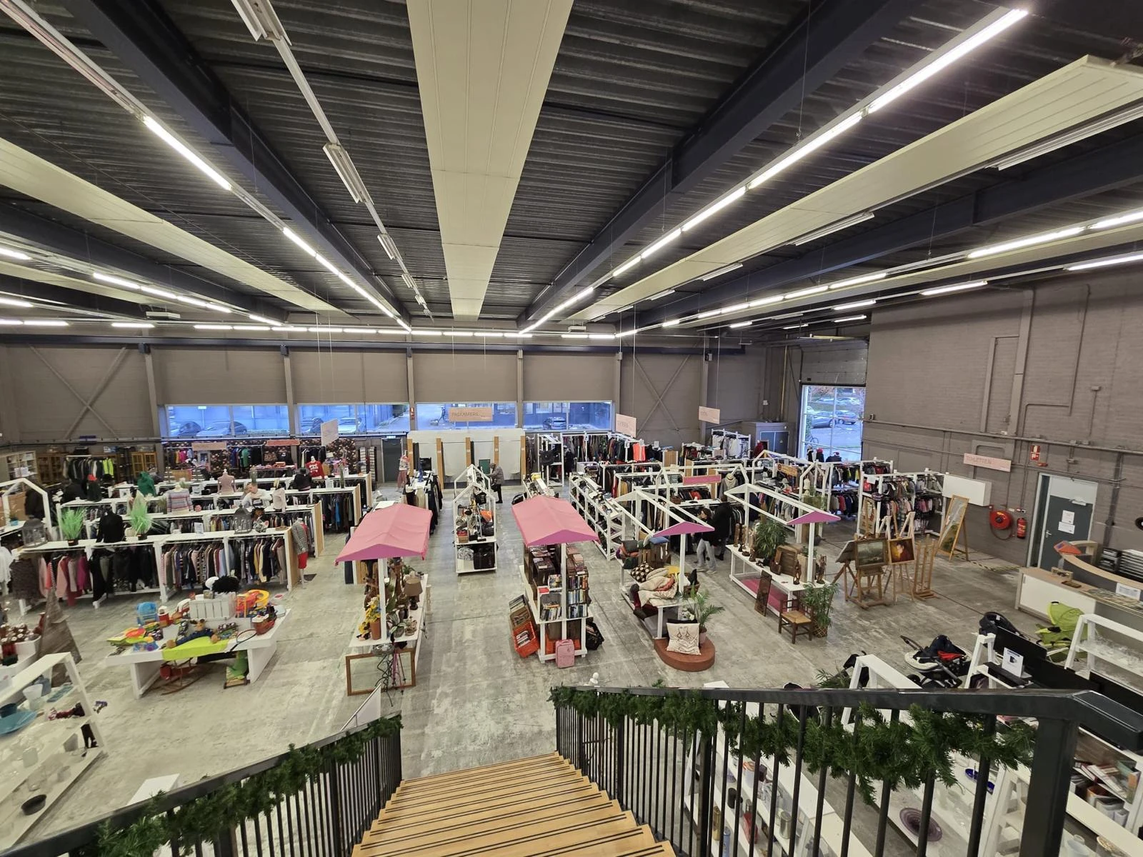 Overzicht van kringloopwinkel met kledingrekken onder roze parasols en verlichte plafondkap