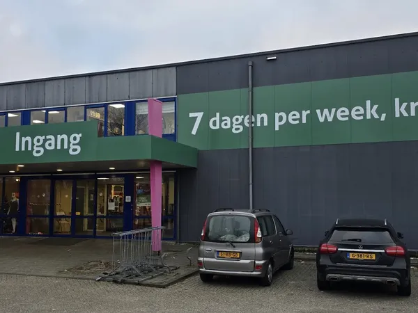 Gevel van kringloopwinkel met groene en donkergrijze bekleding, ingang zichtbaar, twee auto's op parkeerplaats