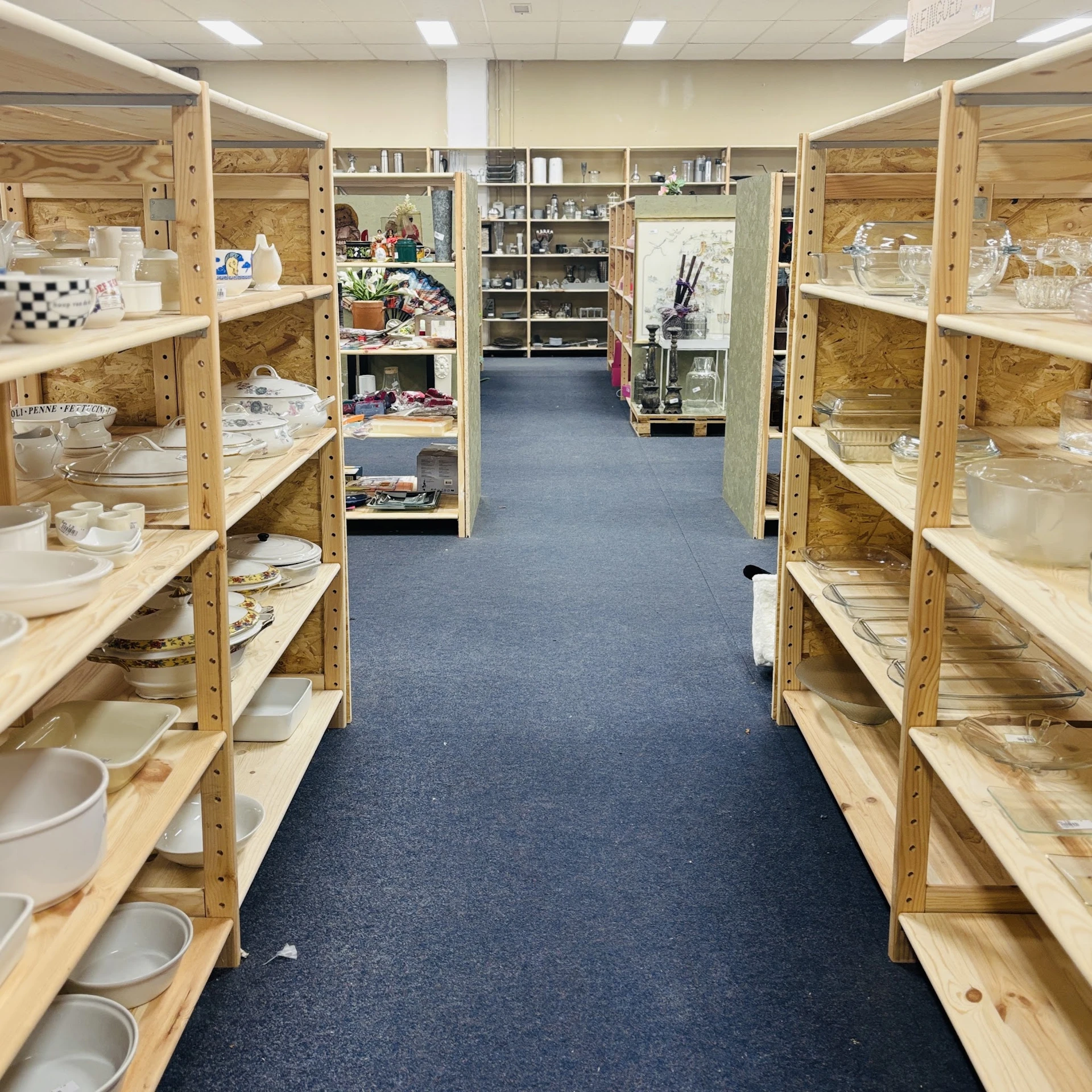 Lange gang in kringloopwinkel met houten stellingen vol serviesgoed en huishoudartikelen aan beide zijden