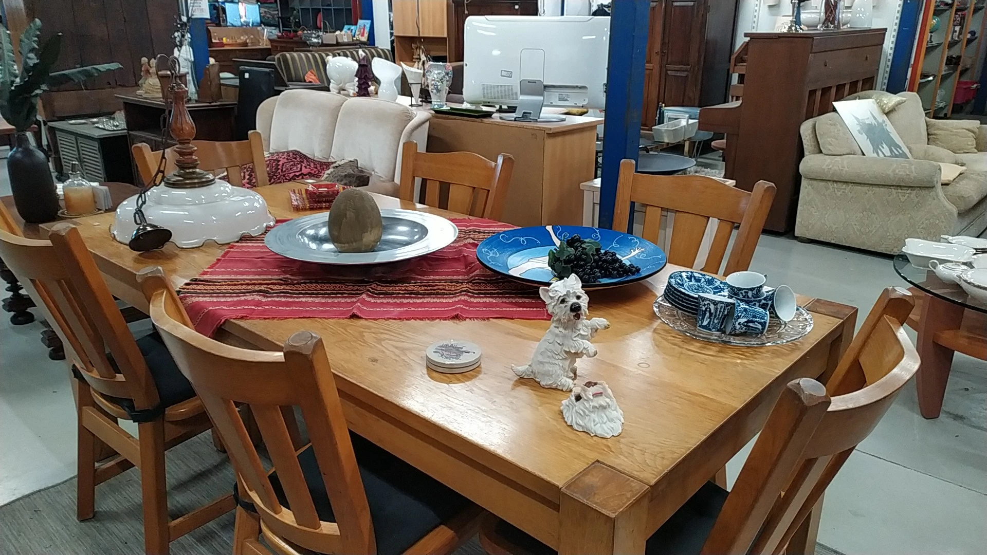 Houten eettafel met stoelen, rode tafelloper en diverse voorwerpen in een kringloopwinkel