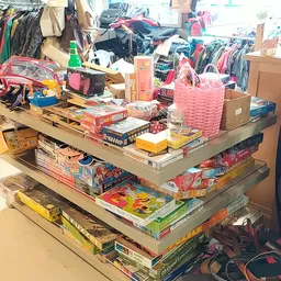 Stapelplateau met speelgoed, bordspellen en kinderartikelen, omgeven door kleding en andere tweedehands goederen